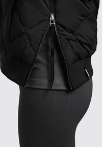 Čierna prešívaná puffer bunda so zipsom a žebrovanými manžetami. Pod ňou je tmavošedý priliehajúci top, skombinovaný s textúrovanými čiernymi legínami.