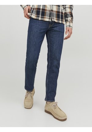 Mężczyzna w niebieskich dżinsach, beżowych zamszowych butach sznurowanych i beżowo-czarno-białej koszuli w kratę na jasnym tle.