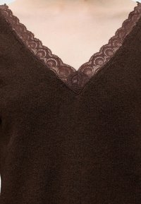 Gros plan montrant un pull en maille marron avec un col en V et une bordure en dentelle marron foncé le long de l'encolure.