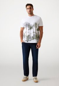 Hvid grafisk t-shirt med print af palmer og bjerge, parret med mørkeblå bukser og beige sneakers med grønne accenter.