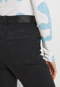 Svart denimjeans med hög midja, med en bakficka och en märkesläderpatch vid midjan. Texturen verkar något grov.