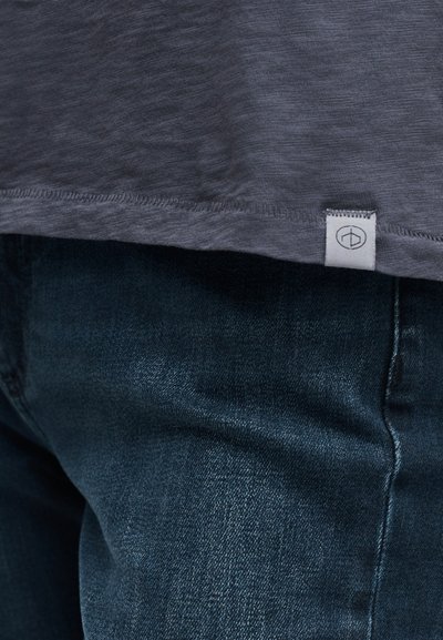 Κοντινή λήψη σκούρου μπλε τζιν τζιν με το τελείωμα ενός γκρι πουκαμίσου που διαθέτει μια μικρή λευκή ετικέτα με μινιμαλιστικό λογότυπο.
