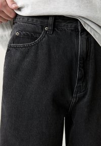 Vaqueros de mezclilla negros con un diseño clásico de cinco bolsillos, costuras resistentes y acentos de metal. Tejido texturizado con un ligero desgaste.