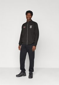 Black zip-up jacket with logo on left chest, paired with black sweatpants and sporty shoes. Smooth fabric, relaxed fit, tucked at waist.