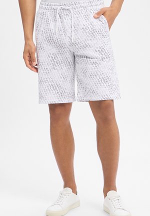 Mężczyzna w białych szortach z sznurkiem i jasnoszarym wzorem powtarzającego się tekstu oraz białych tenisówkach, stojący z jedną ręką w kieszeni.