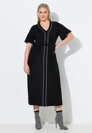 Femme portant une robe midi noire avec un passepoil vertical blanc, des manches courtes et des bottines aux motifs géométriques noir et blanc, debout avec la main sur la hanche.