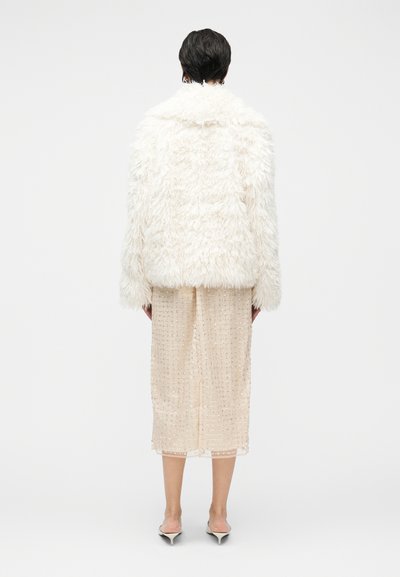 Bela jakna iz umetnega krzna z ohlapnim krojem, nošena čez prosojno kremo obleko z bleščicami, kombinirana s čevlji mule z ostrim nosom.