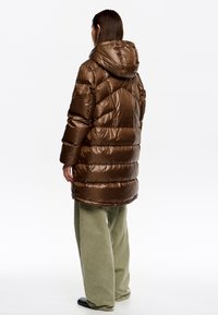 Puchowy brązowy płaszcz z połyskującą fakturą i kapturem, z ukośnym przeszyciem. W zestawie z luźnymi, oliwkowozielonymi spodniami sztruksowymi.