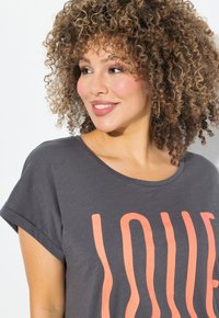 Camiseta gris de manga corta con la palabra "AMOR" en coral en un diseño atrevido. Textura suave, corte relajado, con un escote redondo y mangas ligeramente enrolladas.