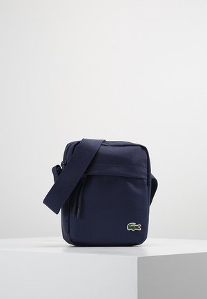 Petit sac bandoulière bleu marine avec sangle réglable et logo crocodile Lacoste sur la poche avant, exposé sur un socle blanc.