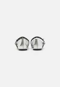 Chaussures métalliques argentées sans dos avec une bride noire à la cheville, présentant des découpes circulaires et un design à bout arrondi. Talon plat, texture lisse.