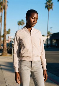 Fiatal fekete nő világos bézs dzsekiben és szürke farmerral napos városi járdán áll, háttérben pálmafákkal és kék éggel.