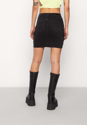 Vrouw die een zwarte denim minirok draagt met achterzakken en zwarte plateau-kniehoge laarzen, staand tegen een effen achtergrond.