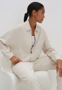 Beige Zip-Jacke mit Reißverschluss und passender Hose aus glattem Stoff, mit Kragen, seitlichen Taschen und legerem Schnitt.