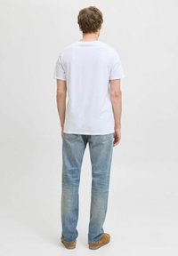 Hombre vestido con una camiseta blanca de manga corta, jeans azules claros y zapatos marrones, de pie de espaldas contra un fondo liso.