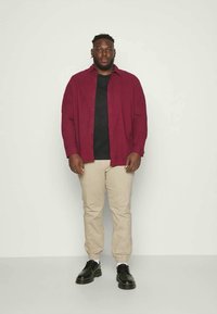 Camicia bordeaux con bottoni sopra una t-shirt nera, abbinata a pantaloni beige e scarpe nere. Tessuto liscio, vestibilità comoda e design semplice.