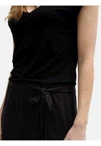Top nero a scollo a V abbinato a pantaloni neri con una cintura annodata. Il tessuto appare morbido con una texture liscia. Tono della pelle visibile sul braccio.
