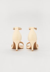 Beige sandály na vysokém podpatku s pásky kolem kotníků, otevřenou zadní částí a úzkým podpatkem. Hladký materiál s matným povrchem a minimalistickým designem.
