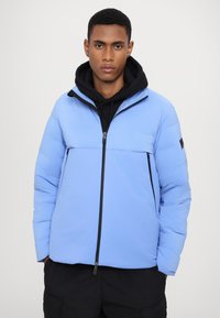 Blue puffer jacket with a black zipper and panel detailing, featuring a hood, long sleeves, and fleece lining. Worn over a black hoodie.