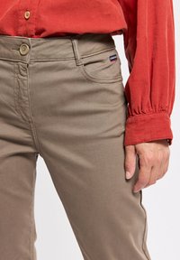 Chinos beiges à coupe slim, fabriqués en tissu de coton doux. Équipés d'une fermeture à bouton, de poches avant et d'une petite étiquette de marque sur le côté.