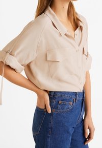 Camisa beige de manga corta con cuello, dos bolsillos en el pecho y mangas enrolladas, combinada con unos pantalones vaqueros azul de cintura alta. Textura de tela suave.