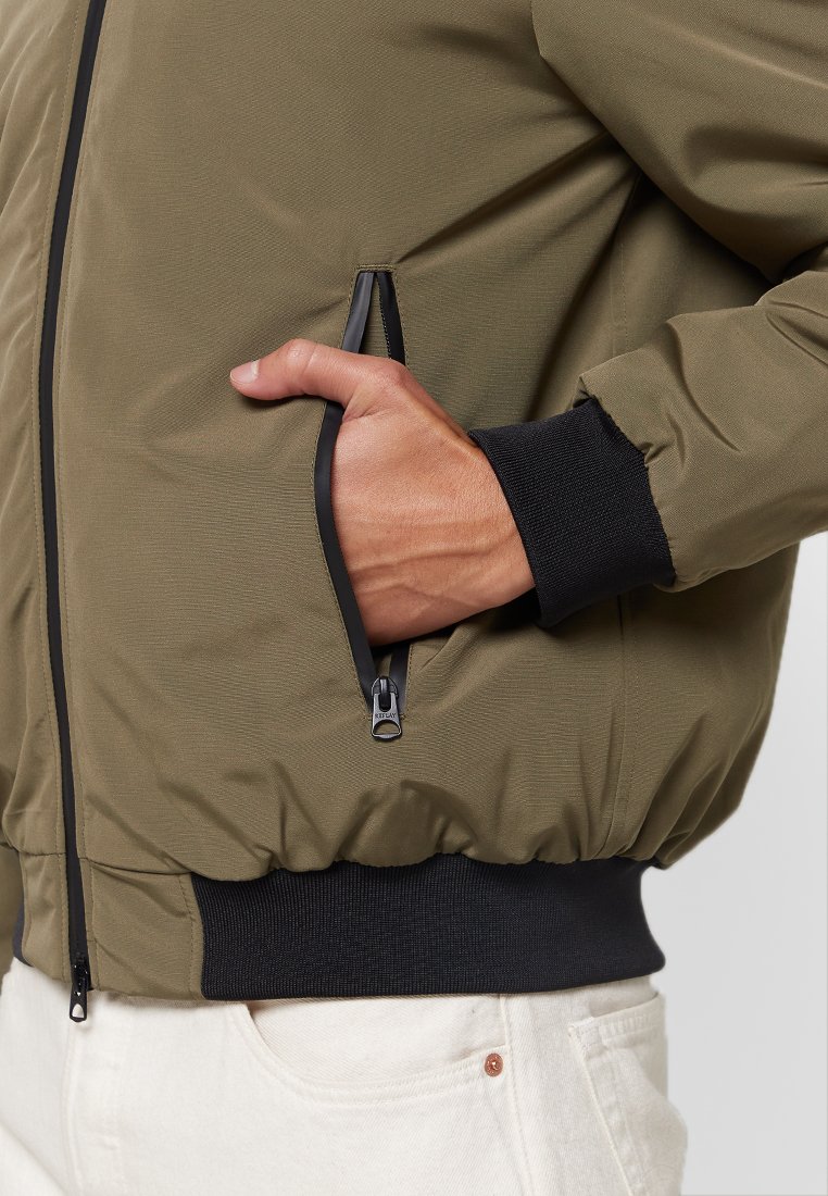 Giacca bomber verde oliva con una texture liscia, tasche laterali con zip, rifinitura nera sui polsini e sull'orlo, e un design aderente.