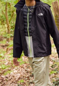 Personne portant une tenue superposée avec une veste noire North Face et un pantalon beige, debout en plein air parmi des arbres et des feuilles tombées.