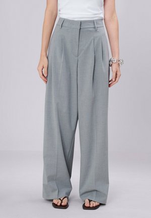 Persoon die wijde lichtgrijze broek draagt, een wit topje, zwarte sandalen en zilveren armbanden aan de rechterpols, staand tegen een effen achtergrond.