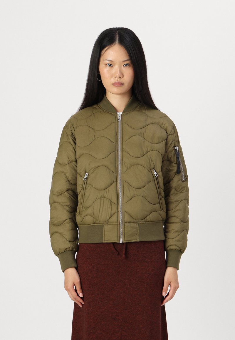 Olive zaļa pūkainā bomber jaka ar viļņu rakstu, ar rāvējslēdzēju priekšā, ribotiem piedurkņu galiem un divām sānu kabatām. Modelis valkā tumši sarkanu svārku zem tās.