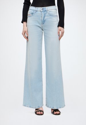 Person iført lyseblå jeans med brede ben, sort langærmet top og sorte sandaler med åbne tæer på en ensfarvet baggrund.