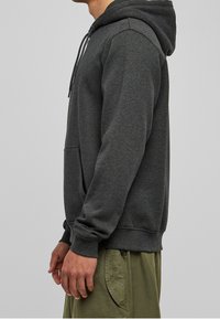 Tumma harmaa huppari, jossa on nyörit, etutasku ja resorit hihansuissa. Kangas näyttää pehmeältä ja rento malli. Päällä vihreiden housujen kanssa.