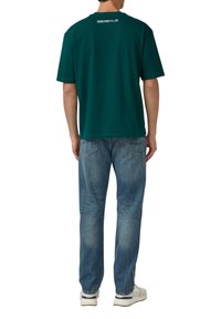 Person wearing a dark green short-sleeve shirt, blue jeans, and white sneakers, standing facing away against a white background.