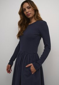 Navy dress with long ribbed sleeves and a fitted bodice, transitioning to a flared skirt with pockets. Smooth fabric and minimalist design.