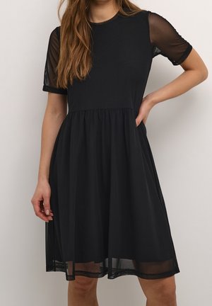 Robe noire longueur genoux avec manches courtes transparentes et taille froncée, portée par une femme aux cheveux longs et ondulés.
