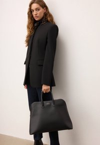 Sort læderhåndtaske med struktureret form, dobbelte håndtag og logo-accent. Model iført en sort blazer og mørke jeans står mod en neutral baggrund.