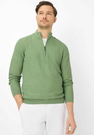 Mand iført en lysegrøn sweater med kvart-lynlås over en hvid skjorte og hvide bukser, stående med den ene hånd i lommen.