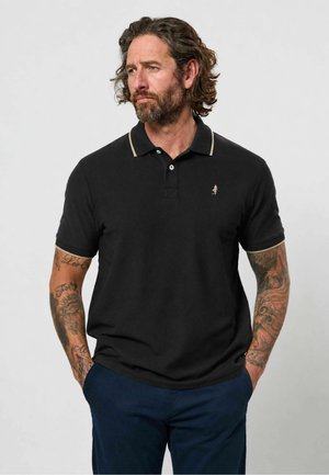 Homme aux cheveux ondulés et tatouages portant un polo noir et un pantalon bleu marine, debout les mains dans les poches, regardant sur le côté.