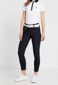 Kvinna iförd slanka svarta byxor, vit bälte, vit polo t-shirt med svart krage och vita sneakers, stående mot en neutral bakgrund.