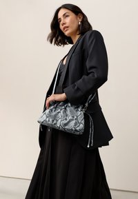 Sac à main imprimé serpent gris et noir avec un haut froncé, une sangle réglable et des accessoires en métal argenté, tenu par une personne portant un blazer noir.