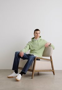 Sudadera verde claro con un logo en el frente, combinada con jeans oscuros y zapatillas blancas, sentado en una silla de marco de madera con tapicería beige.