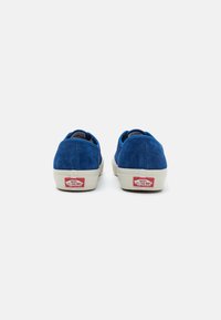 Blue suede sneakers with a low-top design, white rubber sole, and red logo accents on the heel. Smooth texture with a clean finish.