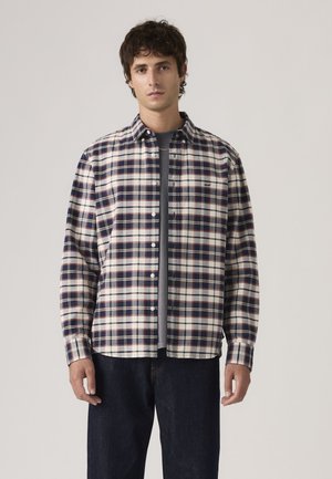 Mann trägt ein navyblaues, weißes und beiges kariertes Hemd über einem grauen T-Shirt mit dunkelblauer Jeans vor neutralem Hintergrund.