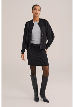 Bomber jacket a righe nere, t-shirt grigia aderente, gonna a righe nere e stivali neri con tacco al ginocchio. Tessuto liscio e vestibilità sartoriale.