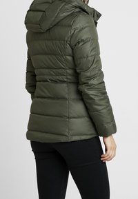 Veste matelassée vert olive avec capuche, présentant un design matelassé, un tissu texturé et une silhouette ajustée. Agrémentée de poignets ajustés.