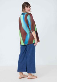 Multicolored blouse in shades of brown, blue, and green with a loose fit and short sleeves, paired with wide-leg blue denim pants and sandals.