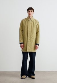 Νεαρός άνδρας που στέκεται μέσα σε εσωτερικό χώρο, φοράει μπεζ παλτό με κουμπιά, μαύρο παντελόνι με καμπάνα και μπεζ παπούτσια, μπροστά σε απλή λευκή τοίχο.