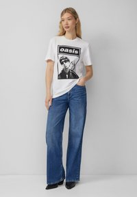White cotton t-shirt featuring a black graphic print of a musician. Paired with wide-leg blue denim jeans and black ankle boots.