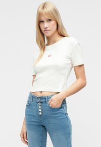 Witte cropped t-shirt met korte mouwen en een subtiele geribbelde textuur, voorzien van een kleine rode hartprint op de borst. Gecombineerd met een spijkerbroek met hoge taille.