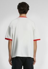T-shirt bianco a maniche corte con dettagli rossi sulle maniche e sul collo. Presenta una vestibilità ampia e un tessuto morbido dalla texture liscia.