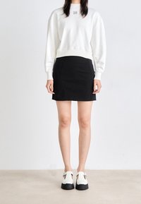 Mujer vestida con una blusa blanca de manga larga, falda corta negra y zapatos de plataforma blancos con suelas negras, de pie frente a un fondo liso.